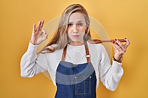 Young blonde woman wearing apron tasting food holding wooden spoon depressed and worry for distress, crying angry and afraid