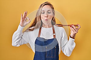 Young blonde woman wearing apron tasting food holding wooden spoon making fish face with mouth and squinting eyes, crazy and