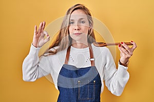Young blonde woman wearing apron tasting food holding wooden spoon relaxed with serious expression on face