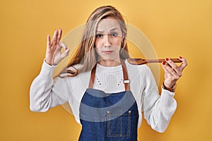 Young blonde woman wearing apron tasting food holding wooden spoon skeptic and nervous, frowning upset because of problem