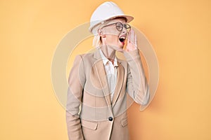 Young blonde woman wearing architect hardhat shouting and screaming loud to side with hand on mouth