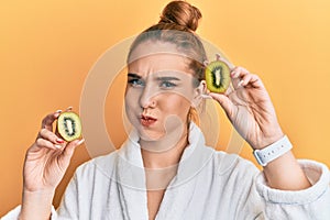 Young blonde woman wearing bathrobe holding fresh kiwi puffing cheeks with funny face