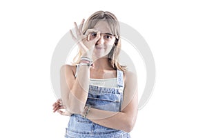Young blonde woman wearing a bib and brace against a white background showing okay sign over eyes