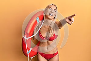 Young blonde woman wearing bikini and holding lifeguard float pointing with finger smiling and laughing hard out loud because