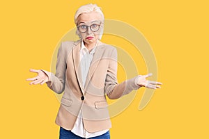 Young blonde woman wearing business clothes and glasses clueless and confused expression with arms and hands raised