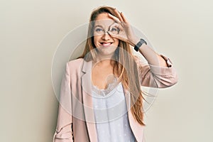 Young blonde woman wearing business jacket and glasses doing ok gesture with hand smiling, eye looking through fingers with happy