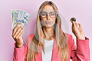 Young blonde woman wearing business style holding dollars and bitcoin looking at the camera blowing a kiss being lovely and sexy