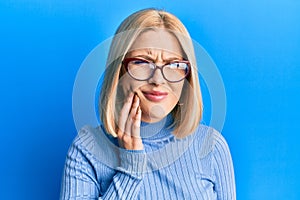 Young blonde woman wearing casual clothes and glasses touching mouth with hand with painful expression because of toothache or
