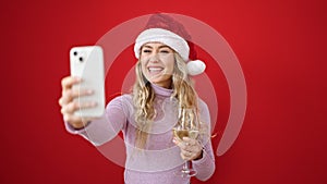 Young blonde woman wearing christmas hat having video call holding glass of wine over isolated red background
