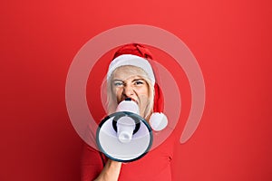 Young blonde woman wearing christmas hat screaming using megaphone over isolated red background