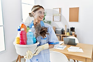 Young blonde woman wearing cleaner uniform holding cleaning products looking away to side with smile on face, natural expression