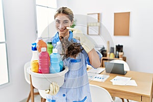 Young blonde woman wearing cleaner uniform holding cleaning products pointing with hand finger to face and nose, smiling cheerful