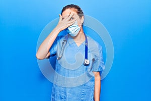 Young blonde woman wearing doctor uniform and medical mask peeking in shock covering face and eyes with hand, looking through