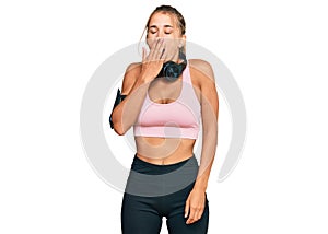 Young blonde woman wearing gym clothes and using headphones bored yawning tired covering mouth with hand