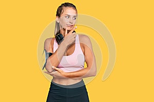 Young blonde woman wearing gym clothes and using headphones with hand on chin thinking about question, pensive expression