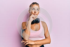 Young blonde woman wearing gym clothes and using headphones serious face thinking about question with hand on chin, thoughtful