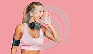 Young blonde woman wearing gym clothes and using headphones shouting and screaming loud to side with hand on mouth
