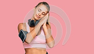 Young blonde woman wearing gym clothes and using headphones sleeping tired dreaming and posing with hands together while smiling