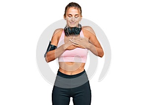 Young blonde woman wearing gym clothes and using headphones smiling with hands on chest with closed eyes and grateful gesture on