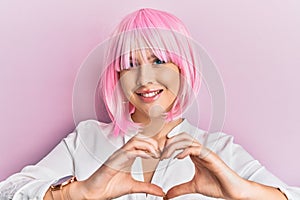 Young blonde woman wearing pink wig smiling in love showing heart symbol and shape with hands