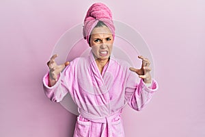 Young blonde woman wearing shower towel cap and bathrobe shouting frustrated with rage, hands trying to strangle, yelling mad