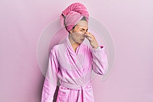 Young blonde woman wearing shower towel cap and bathrobe smelling something stinky and disgusting, intolerable smell, holding