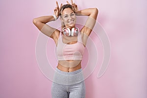 Young blonde woman wearing sportswear and headphones posing funny and crazy with fingers on head as bunny ears, smiling cheerful