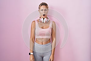 Young blonde woman wearing sportswear and headphones in shock face, looking skeptical and sarcastic, surprised with open mouth