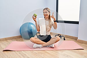 Young blonde woman wearing sportswear and towel holding healthy apple pointing fingers to camera with happy and funny face