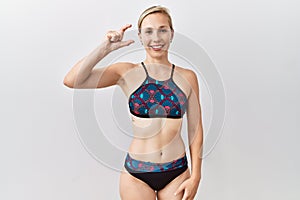 Young blonde woman wearing sporty bikini over isolated background smiling and confident gesturing with hand doing small size sign