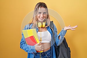 Young blonde woman wearing student backpack and holding books smiling cheerful presenting and pointing with palm of hand looking