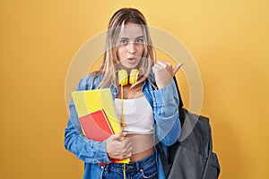 Young blonde woman wearing student backpack and holding books surprised pointing with hand finger to the side, open mouth amazed