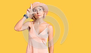 Young blonde woman wearing summer hat making fun of people with fingers on forehead doing loser gesture mocking and insulting
