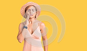 Young blonde woman wearing summer hat touching mouth with hand with painful expression because of toothache or dental illness on