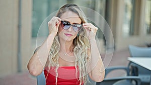 Young blonde woman wearing sunglasses sitting on table smiling at coffee shop terrace