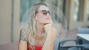 Young blonde woman wearing sunglasses sitting on table smiling at coffee shop terrace