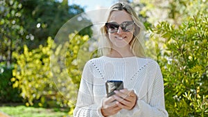 Young blonde woman wearing sunglasses using smartphone at park