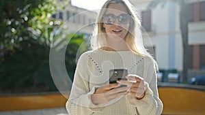 Young blonde woman wearing sunglasses using smartphone at park