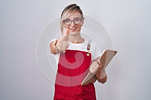 Young blonde woman wearing waiter uniform holding clipboard pointing fingers to camera with happy and funny face