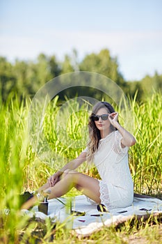 Young blonde woman in white dress and sunglasses is sitting on a blanket in tall grass