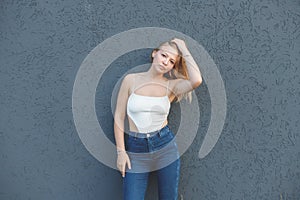 young blonde woman in the white sleeveless t-shirt and blue jeans standing over gray wall background