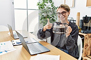 Young blonde woman working at the office holding coffee pot smiling happy and positive, thumb up doing excellent and approval sign