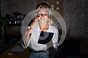 Young blonde woman working at the office at night pointing with hand finger to face and nose, smiling cheerful