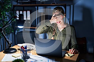 Young blonde woman working at the office at night smiling and laughing with hand on face covering eyes for surprise