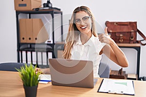 Young blonde woman working at the office wearing glasses smiling happy and positive, thumb up doing excellent and approval sign