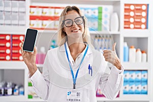 Young blonde woman working at pharmacy drugstore showing smartphone screen smiling happy and positive, thumb up doing excellent