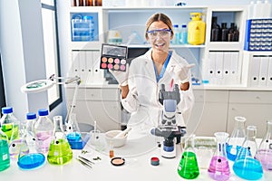 Young blonde woman working at scientist laboratory with make up pointing thumb up to the side smiling happy with open mouth