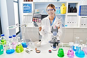 Young blonde woman working at scientist laboratory with make up smiling happy pointing with hand and finger to the side