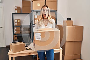 Young blonde woman working at small business ecommerce holding big box afraid and shocked with surprise and amazed expression,