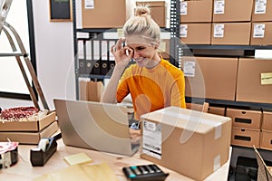 Young blonde woman working at small business ecommerce using laptop smiling happy doing ok sign with hand on eye looking through